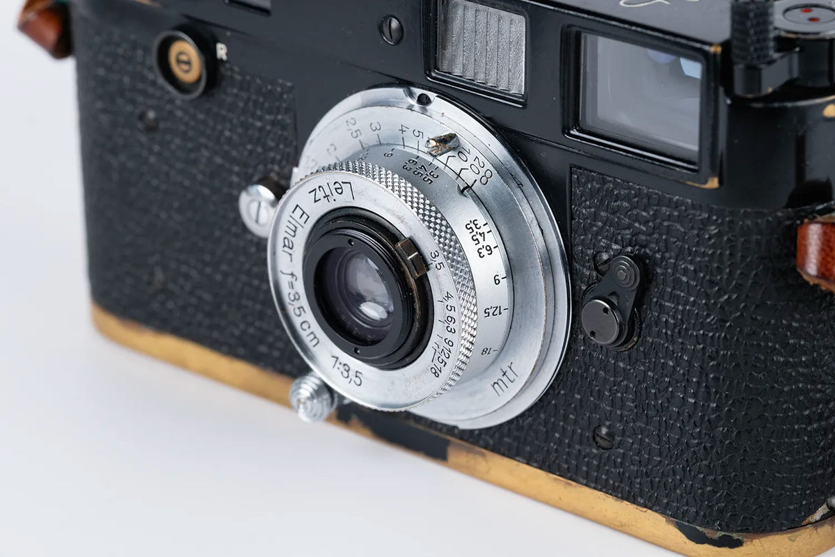 Close-up of a vintage black Leica rangefinder camera body with a silver metal lens and textured leatherette grip, photographed on a white background.