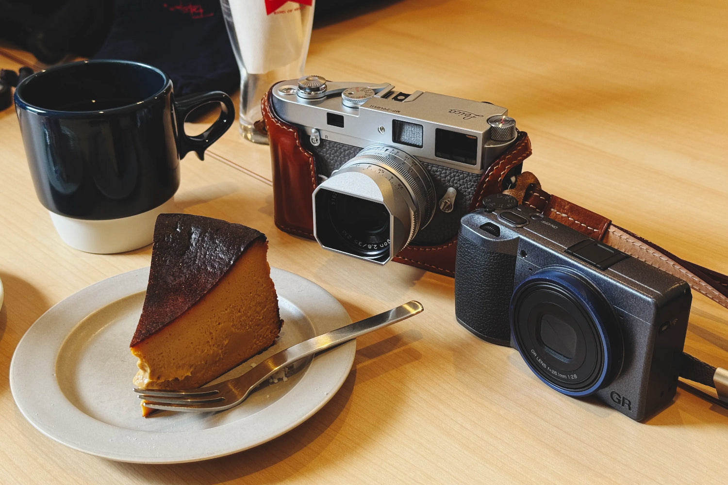Leica camera with soft release button and Ricoh GR on a table with a slice of cake and a cup.